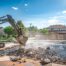 A professional, modern demolition scene in a sunny, upscale Oklahoma City backyard. A large, clean excavator with an F5 Demolition logo is actively breaking up the concrete of an in-ground swimming pool. The background should feature a well-maintained, classic suburban home. The sky is bright blue with a few white clouds. The mood is constructive and informative, focused on the transition from old pool to clean yard.