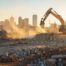 A dynamic, slightly elevated wide shot capturing a complex demolition scene in the background of a modern Oklahoma City skyline (suggesting OKC location). The foreground features heavy equipment—a bright yellow excavator or bulldozer visible—actively breaking a concrete slab or the side of a structure. The scene is clean, emphasizing professionalism and controlled chaos, with the sun setting, casting a golden glow on the dust and machinery.