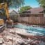 A bright, sunny suburban backyard in Oklahoma City. In the foreground, an older, unused, cracked in-ground swimming pool is being actively demolished by a yellow excavator with a concrete breaker attachment. The machine is actively breaking the bond beam.