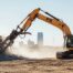 A striking, high-quality, wide-angle photograph of a professional demolition site in Oklahoma City. In the foreground, a large excavator with the F5 Demolition logo visible on the arm, actively breaking up a large section of concrete house foundation, creating dust and action. In the background, a clear view of a dry, slightly dusty Oklahoma skyline or a recognizable local OKC structure (like a modern building or distant transmission tower) under a bright, sunny sky. The atmosphere should feel professional, powerful, and focused on clean site prep.