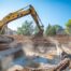 A high-resolution, wide-angle shot of a professional yellow excavator carefully removing a concrete in-ground swimming pool in an Oklahoma City suburban backyard. Bright sunny day, clear blue sky, dust being controlled by a water mist. Professional construction crew in safety vests visible in the background.