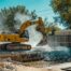 A high-resolution, realistic photo of a professional yellow excavator carefully removing a concrete inground pool in a suburban Oklahoma City backyard. Clear blue sky, dust control water mist visible, professional crew in high-visibility vests nearby. F5 Demolition style branding environment.