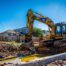 A high-resolution, realistic photo of a professional yellow excavator carefully removing a concrete pool in a suburban Oklahoma City backyard. Bright sunny day, clear blue sky, workers in high-visibility vests and hard hats. Professional branding on the equipment, clean job site.