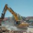 A high-resolution, realistic photo of a professional yellow excavator carefully removing concrete from an old backyard swimming pool in a suburban Oklahoma City neighborhood. Clear blue sky, dust control water spray visible, professional workers in high-visibility vests nearby. 8k resolution, cinematic lighting.